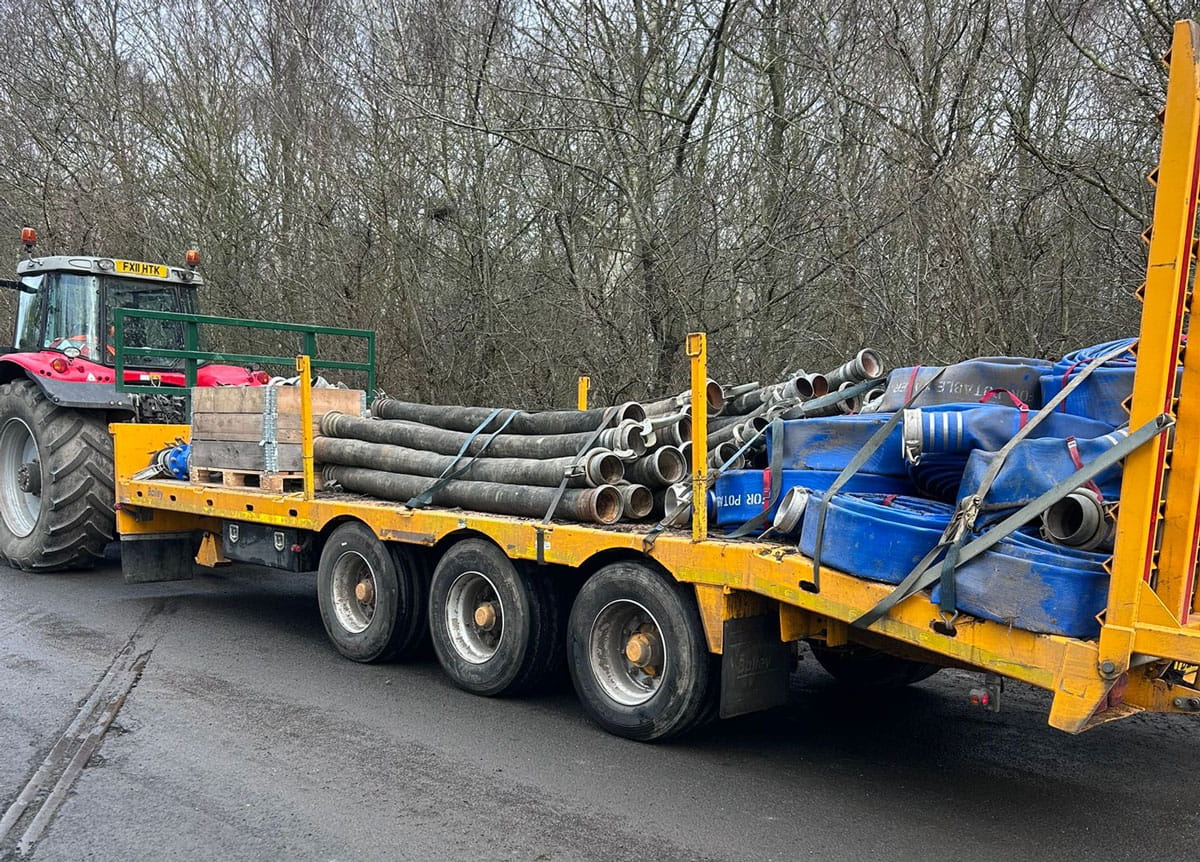 Tractor towing a flatbed trailer loaded with industrial hoses and pipeline equipment for temporary over-pumping operations.